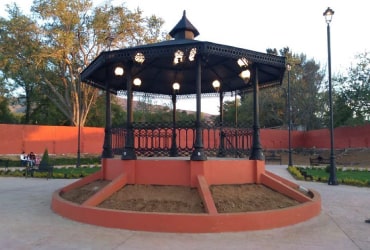 Kiosco colonial octagonal de hierro fundido con barandales decorativos y techo a dos aguas en parque público